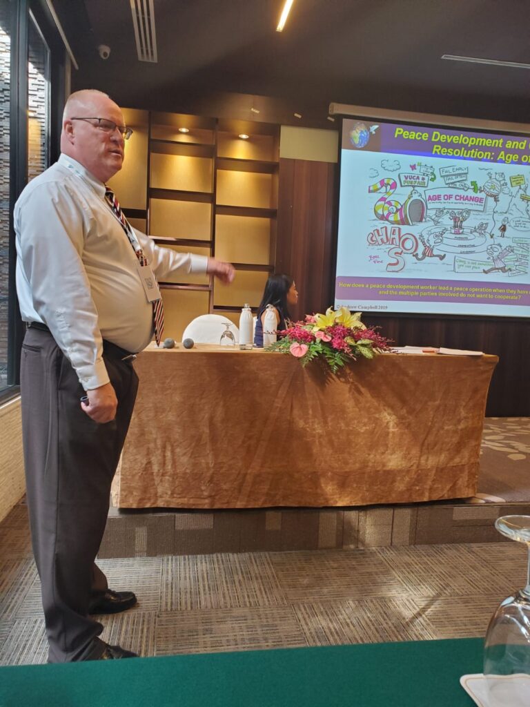 Presenter discusses peace development and resolution concepts during a seminar, with a visual aid displayed on a screen behind.