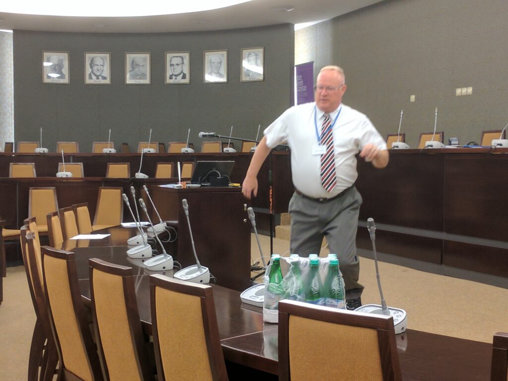 A man in business attire stands near a conference table in a formal meeting room, preparing for an event. Bottled water is visible on the table.