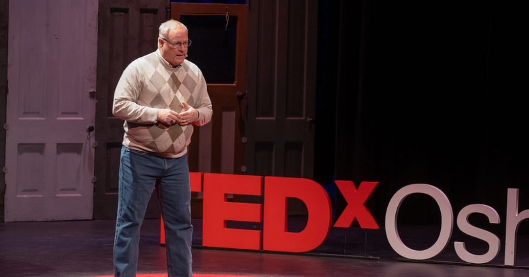 A speaker presents at a TEDx event, standing in front of vibrant red "TEDx" letters against a backdrop of various doors.