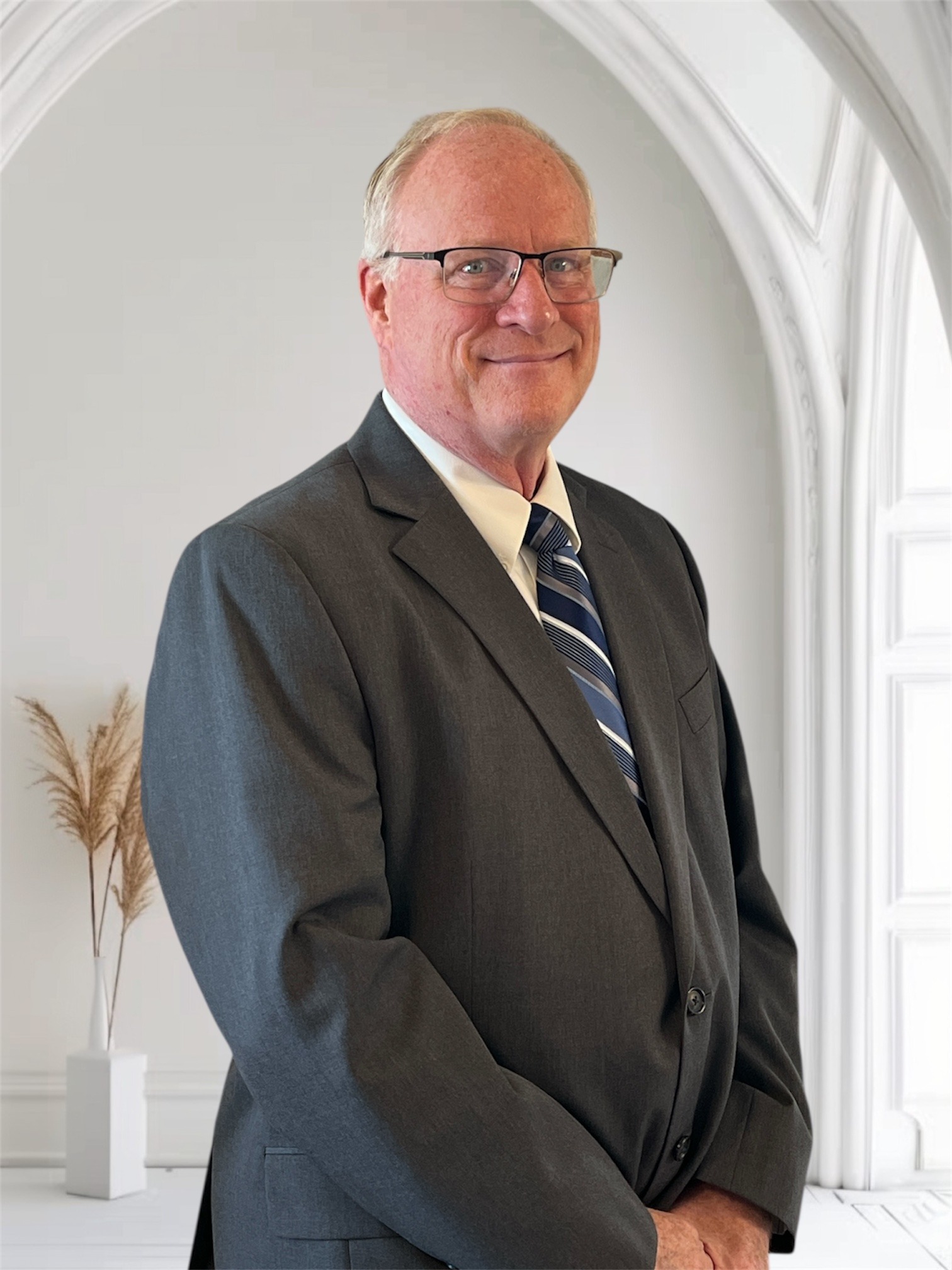 Professional man in a grey suit and glasses smiles confidently against a neutral background, ideal for a business profile or introduction.