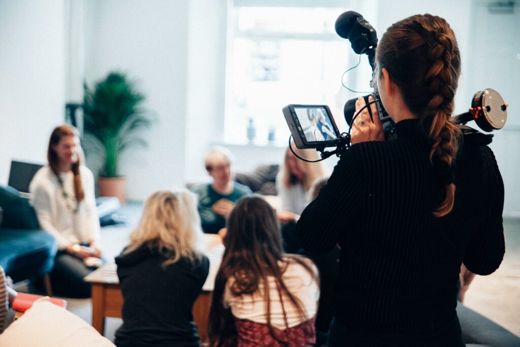 A person with a camera captures a group discussion among women seated around a table, fostering collaboration and creativity in a cozy setting.