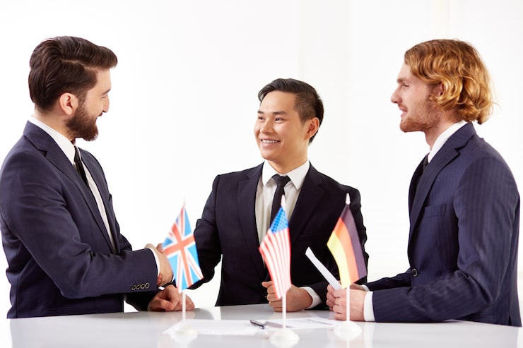 Three men in suits happily shake hands around a table featuring small U.K., U.S., and German flags, suggesting international business collaboration.