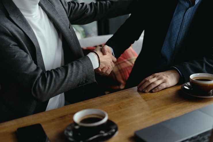 Two people in suits shake hands across a wooden table, with cups of coffee beside them. The scene conveys a professional and amiable meeting.