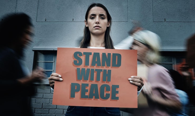 A woman holds a sign reading "Stand with Peace" while people move blurred around her, conveying a message of calm amidst chaos.