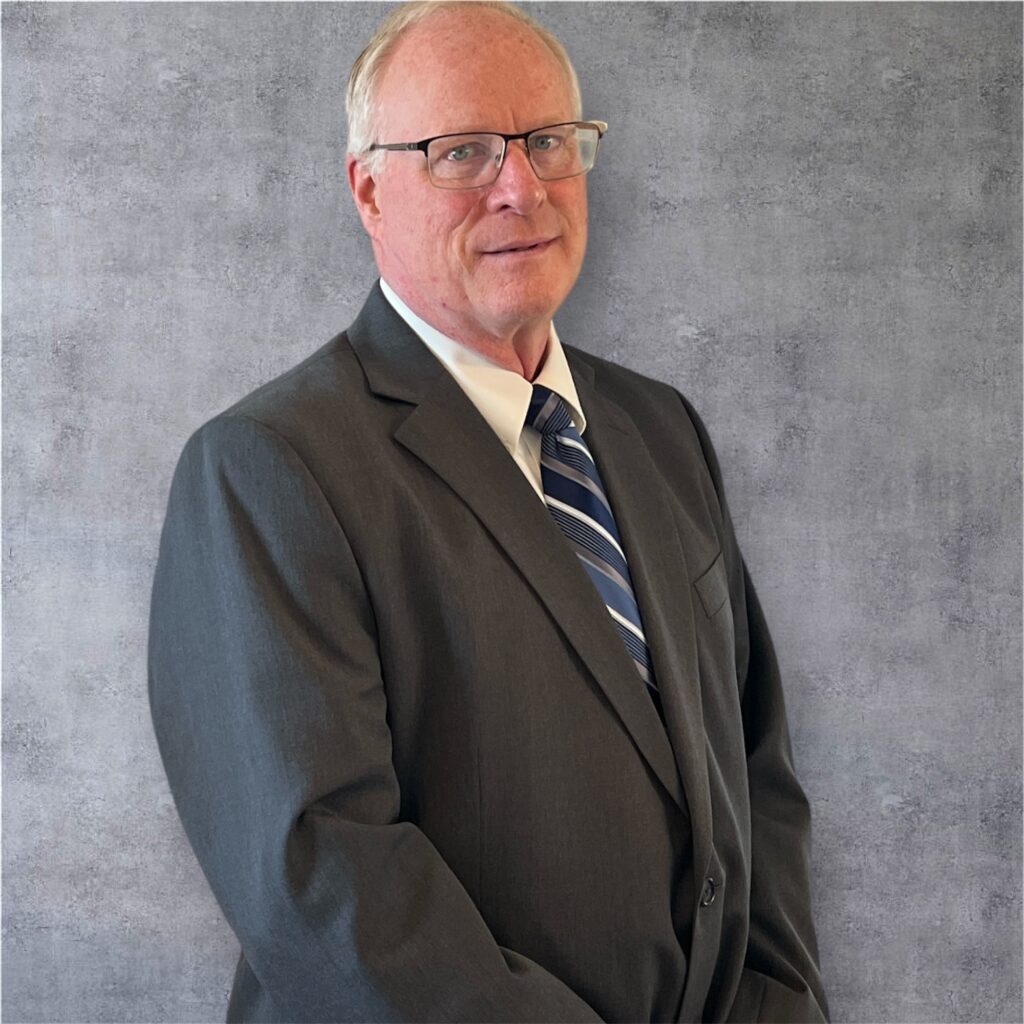 A man in a dark suit with a striped tie stands confidently against a textured gray background, conveying professionalism and approachability.